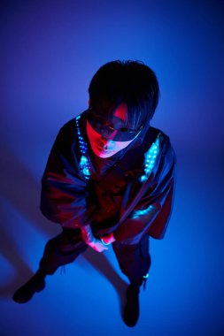 Yakışıklı genç bir adam şık bir pozla duruyor, mavi neon arka planda güneş gözlüğü takıyor..