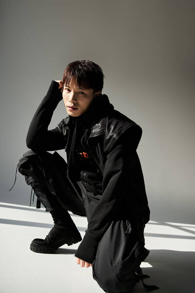 Stylish Asian man crouches in a modern studio, showcasing charisma and fashion sense.
