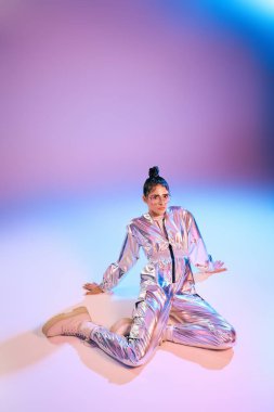 Metalik bir elbise giyen genç bir kadın renkli bir arkaplan karşısında şık bir şekilde dizlerinin üzerinde poz veriyor..