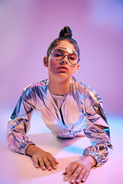 A young woman leans over a surface in a shiny holographic outfit amid vibrant colors.