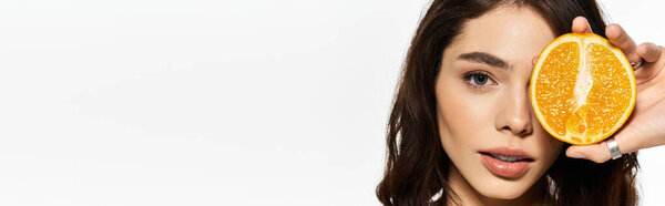 Stylish young woman enjoys summer vibes, playfully posing with a fresh orange slice.