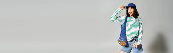 Уверенно позирующая молодая женщина в шикарной летней одежде и игривой кепке на простом фоне.