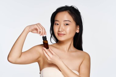 Kendine güveni tam genç bir kadın, elinde küçük bir güzellik serumu şişesiyle temiz bir arkaplana gülümsüyor..
