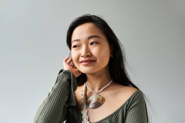 Genç Asyalı kadın saçlarına dokunurken ve zarif kolyesini gösterirken gülümsüyor..