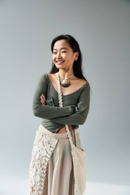 Genç bir Asyalı kadın kollarını kavuşturup minimalist bir alanda sevinçle gülümsüyor..