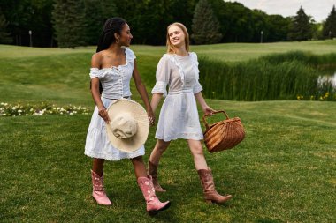 İki genç kadın çimenli bir parkta el ele yürüyorlar, yazlık elbiseler giyiyorlar..