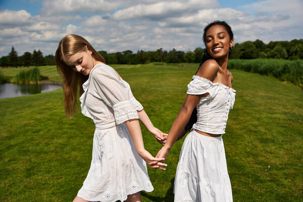 A beautiful couple of young women hold hands while walking on lush grass under a bright sky.