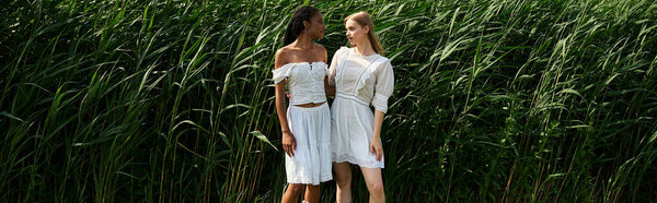Two beautiful women stand together in a vibrant green field, sharing smiles and laughter.