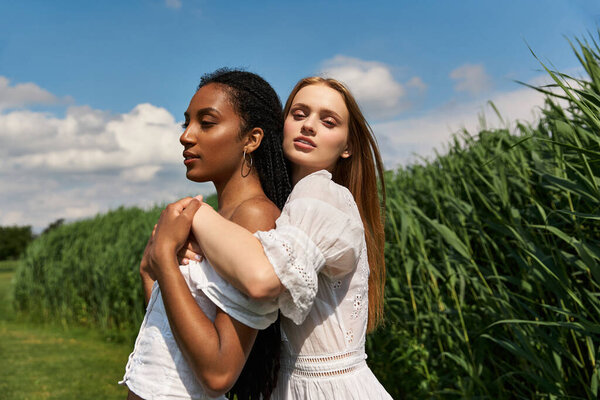 Two young women share a joyful moment in a lush green field beneath a clear blue sky.