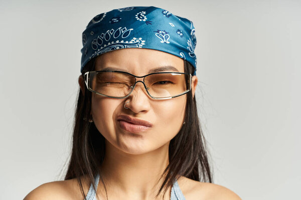 Young woman with a blue bandana and glasses shows a playful expression on a neutral background.