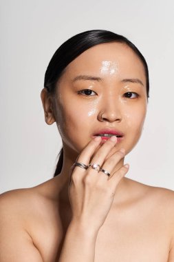 Genç bir Asyalı kadın nazikçe yüzüne dokunuyor, pürüzsüz cildini ve şık yüzüklerini gösteriyor..
