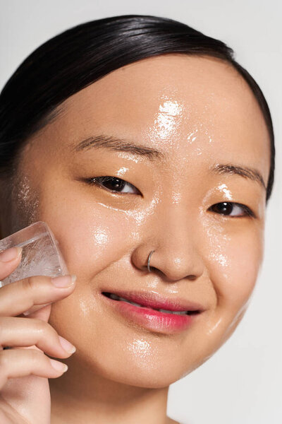 A young Asian woman smiles as she applies ice to her glowing skin, enhancing her beauty.