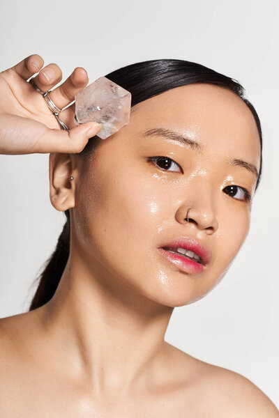 A young beautiful asian woman holds an ice cube to her glowing skin while pampering herself.