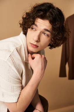 Kıvırcık saçlı genç adam, tarafsız bir arkaplan karşısında hiç çaba harcamadan moda bir yaz kıyafeti içinde poz veriyor..