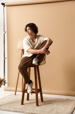 Kıvırcık saçlı, güzel bir genç adam, yazlık kıyafetinin altını çizerek, bir tabureye poz veriyor..