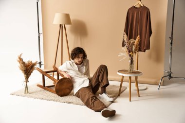 Kıvırcık saçlı genç bir adam minimalist bir stüdyo ortamında son moda bir yaz kıyafeti giyiyor..