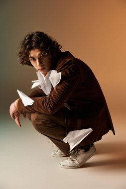 Kahverengi kadife ceketli kıvırcık saçlı genç adam şık bir şekilde çömelmiş, etrafı kağıt uçaklarla çevrili..