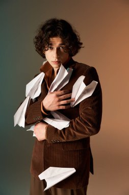 Kıvırcık saçlı genç adam, ceketinde kağıt uçaklar olan bir stüdyoda güven içinde poz veriyor..