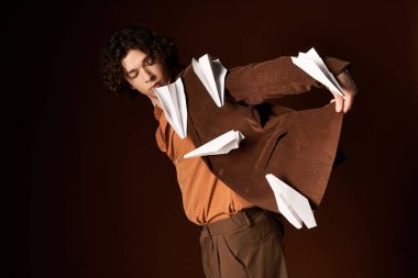 Kahverengi kadife ceketli genç bir kıvırcık adam kağıt uçaklar sergiliyor, yaratıcılığı somutlaştırıyor..