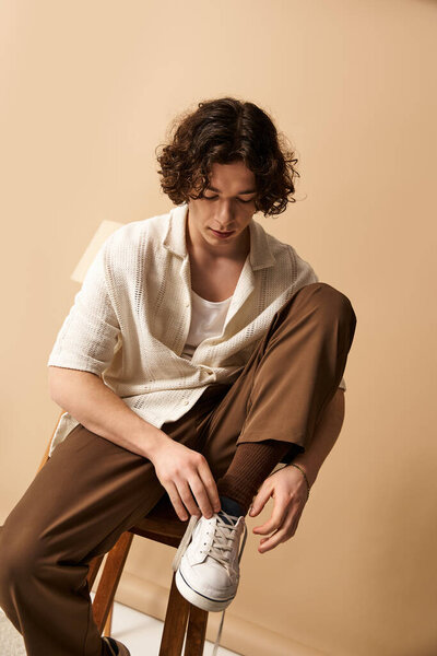 A good looking young man with curly hair is adjusting his stylish shoes while seated in a studio.