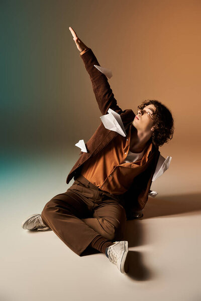 A young man sits gracefully, reaching for the sky amid floating paper planes in a studio.