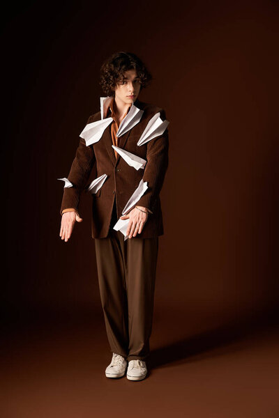 A stylish young man in a brown blazer with paper planes poses confidently in a studio.