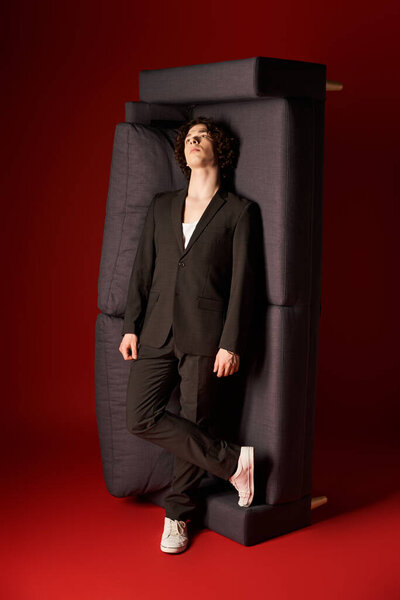 Красивый молодой человек в стильной черной одежде уверенно стоит на диване и красном фоне.