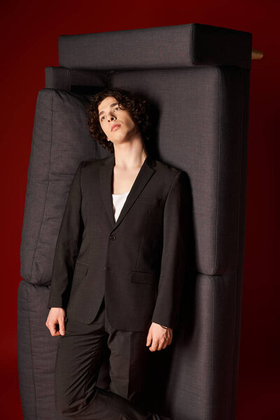 A young man in black attire leans against modern couch in a vibrant studio setting.