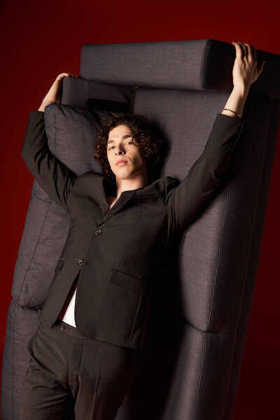 A stylish young man with curly hair poses confidently against a bold red background with couch