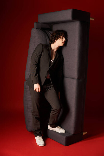 Красивый молодой человек в черном стильном наряде творчески позирует с диваном в студии.