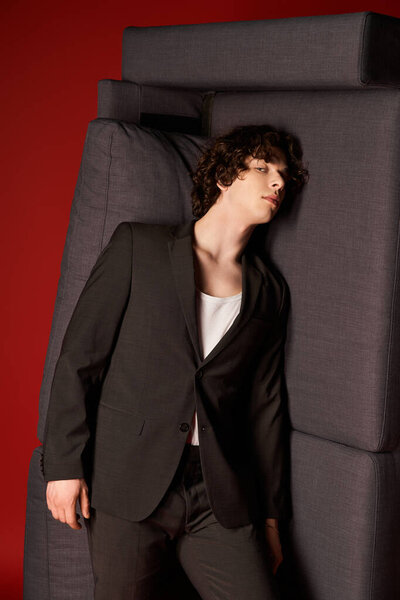 A young man with curly hair leans on grey cushions, flaunting his stylish black outfit.