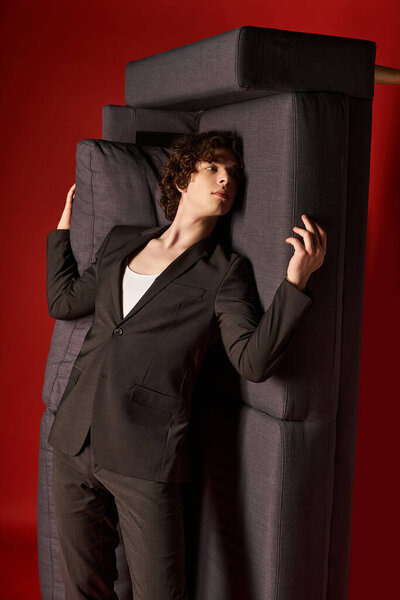 A curly-haired young man models sleek black attire, artistically engaging with his surroundings.