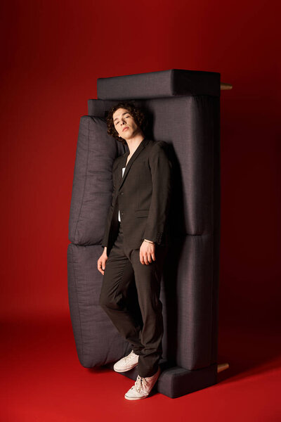 A young man with curly hair poses confidently in a sleek black outfit against a red backdrop.