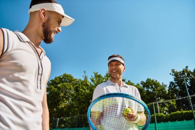 İki mutlu genç adam güneşli bir kortta tenis oynuyorlar, birlikte gülüyorlar ve eğleniyorlar..