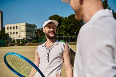 İki adam yaz güneşinin altında neşeyle tenis oynuyorlar, kahkahalarını paylaşıyorlar..