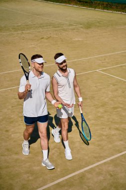 Güneşli bir tenis kortunda iki adam birlikte yürüyor ve gülüyor, eğlenceli bir tenis oyunu oynuyorlar..