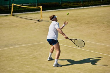 Beyaz spor giyimli yetenekli bir oyuncu güneşli bir kortta tenis topuna vurmaya hazırlanıyor..