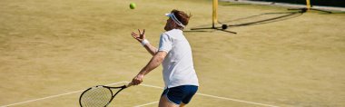 Beyazlar içindeki genç bir adam güneşli bir kortta tenis topuna hizmet etmeye hazırlanıyor.