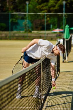 Aydınlık bir yaz günü, beyaz giysili bir adam tenis antrenmanı sırasında bir topu almak için eğilir..