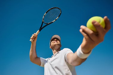 Beyaz spor giyimli bir adam, parlak bir yaz gökyüzünün altında tenis topuna hizmet eder..