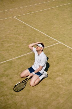 Beyaz spor kıyafetleri giymiş bir adam tenis kortunda diz çöküp yaz havasını içine çekiyor..