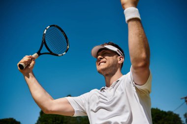 Beyaz spor kıyafetli yakışıklı bir adam açık bir yaz gökyüzü altında tenis zaferini kutluyor..