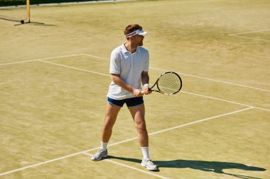 Beyaz giysili genç bir adam yaz boyunca güneşli bir tenis kortunda hizmet etmeye hazırlanıyor..