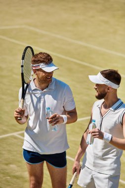 İki genç adam tenis kortunda kahkahalarını paylaşırken su şişeleriyle ferahlatıyorlar..