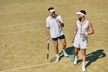 İki yakışıklı adam yaz günü tenis oynadıktan sonra rahatlıyor, suyu yudumluyor ve birlikte gülüyorlar..