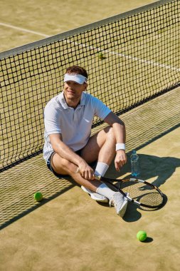 Beyaz spor giysili atletik bir adam tenis kortunda güneşlenmek için biraz zaman ayırıyor..