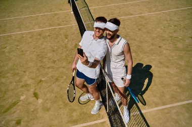 Yaz boyunca güneşli bir tenis kortunda iki genç adam gülümseyip selfie çekiyorlar..