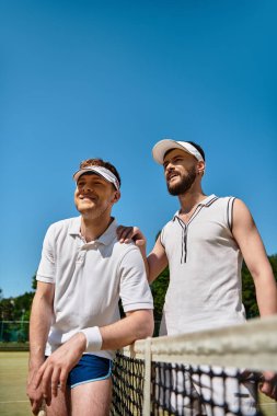 Tenis kortundaki iki adam, yaz güneşinde maçın keyfini çıkarırken gülümsüyor ve gülüyorlar..
