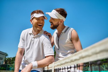 Parlak yaz gökyüzü altında tenis oynarken iki adam gülüşüyor, neşe ve sevgi sergiliyorlar..