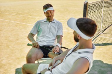 Güneşli bir günde tenis maçından sonra dinlenen iki adam, canlı bir atmosferde bağlantılarını gösteriyorlar..